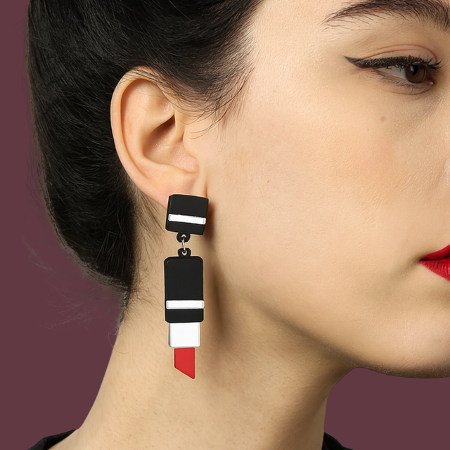 visage féminin sur fond prune portant les boucles d'oreilles en acrylique découpé au laser en forme de tube de rouge à lèvre. la jeune femme porte du rouge à lèvre et elle est de profil.