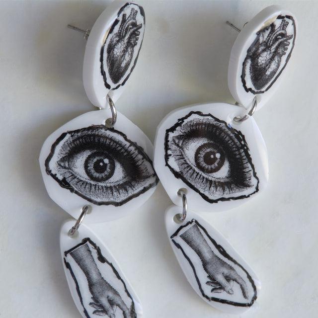 Gros plan sur les motifs cœur anatomique, œil et main des boucles d’oreilles DRAMARAMA