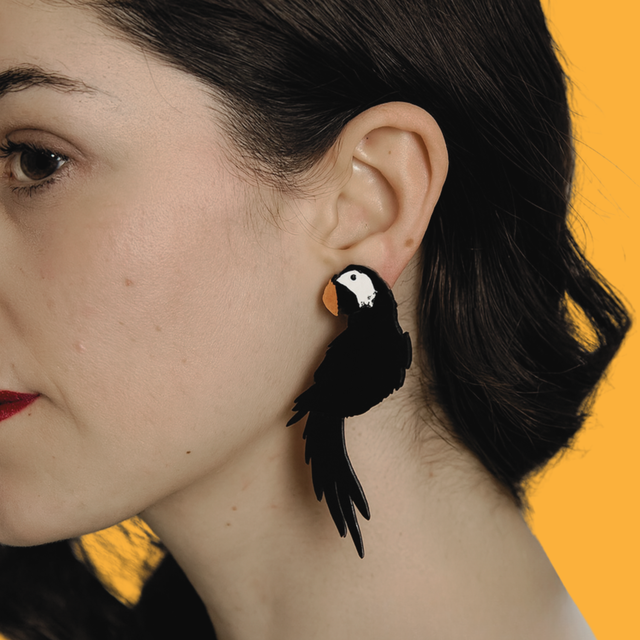 modèle féminin posant de profil, elle porte les boucles d'oreilles en acrylique noir découpé au laser en forme de perroquet sur un fond jaune.