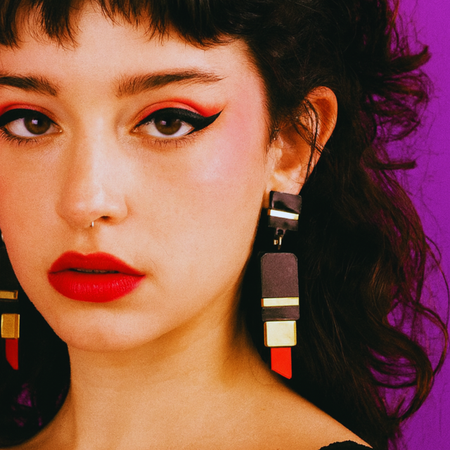 modèle féminin sur fond violet portant les boucles d'oreilles en acrylique découpé au laser en forme de tube de rouge à lèvres.