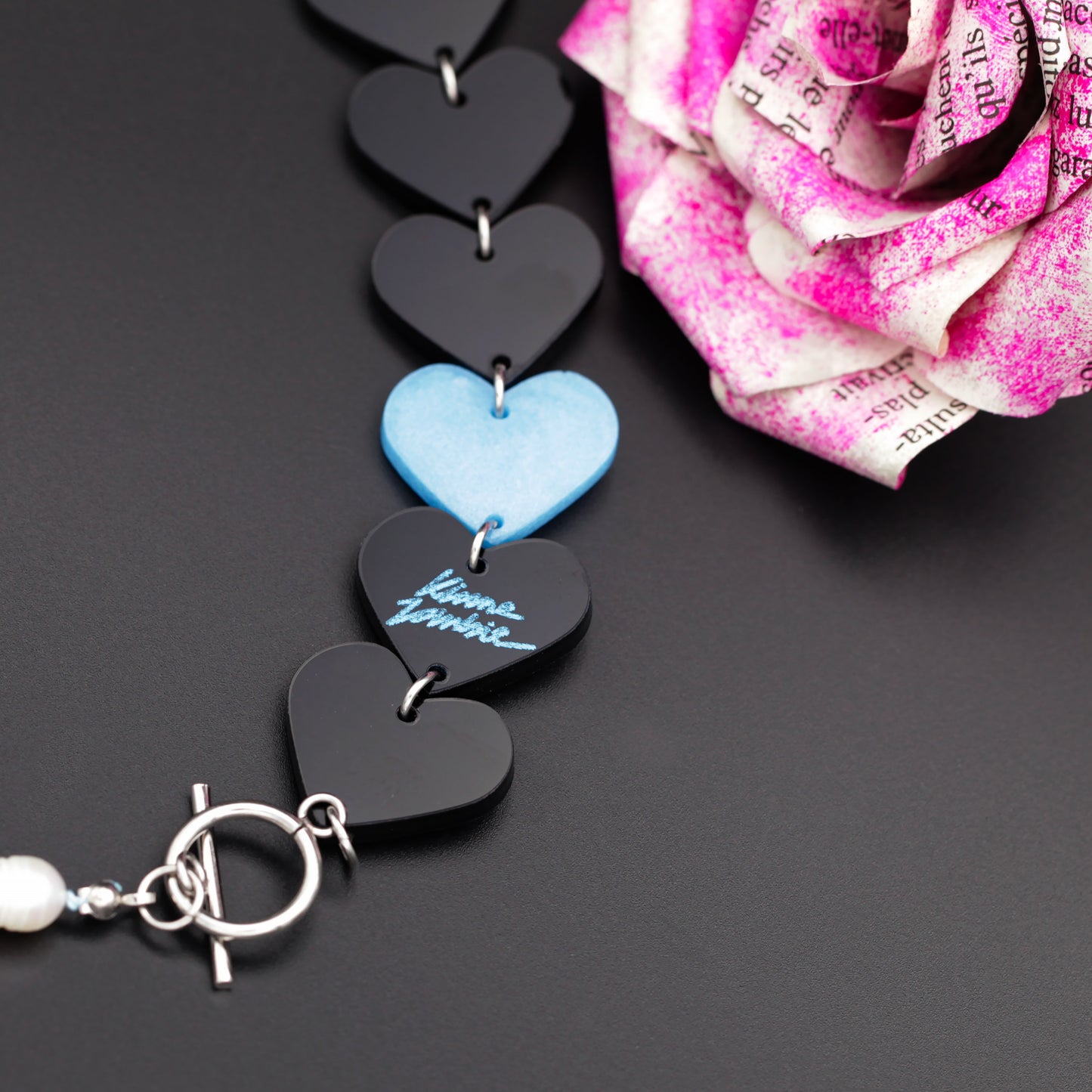 Close-up of a laser-cut and resin black and blue hearts necklace with freshwater pearls and toggle clasp with a newspaper flower in the back on a black background. 