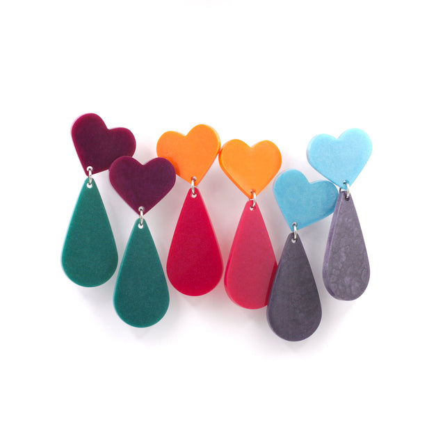 This is a picture of three pairs of earrings of the same design. The top is a heart and the pendant a teardrop. They are from three different colors. on the left, the heart are purple and green teardrop, at the center, orange hearts with pink teardrops and at the right side, blue heart with purple teardrop. They are hanging from a white background.