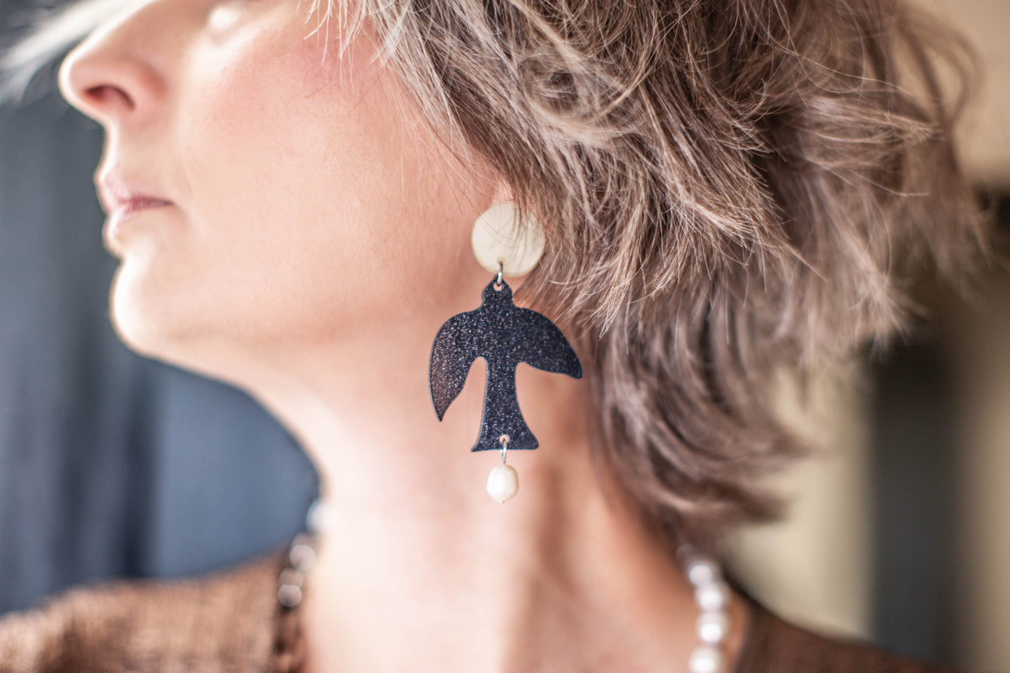 this is a picture of a woman profile we can't see the top of her face. she is wearing statement clip on earrings composed by a white dot (moon) and black glitter bird and a freshwater pearl. the background is blue and white.