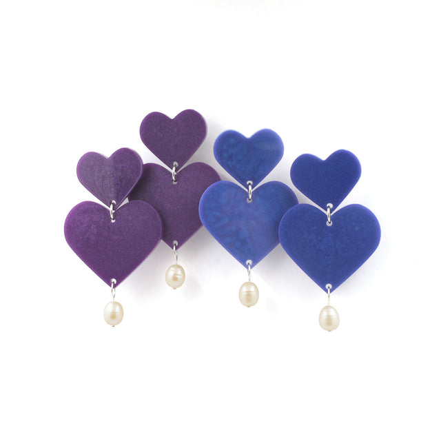 this is a picture of two pairs of earrings of the same design but different colors. the top of the earrings are hearts and the pendant are bigger hearts with freshwater pearls hanging from them. The one one the left are purple earrings and the one on the right are blue earrings. They are on a white background.