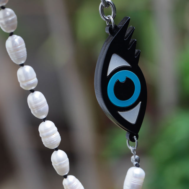 Close-up of a laser-cut evil eye and freshwater pearls necklace on a vegetal background.