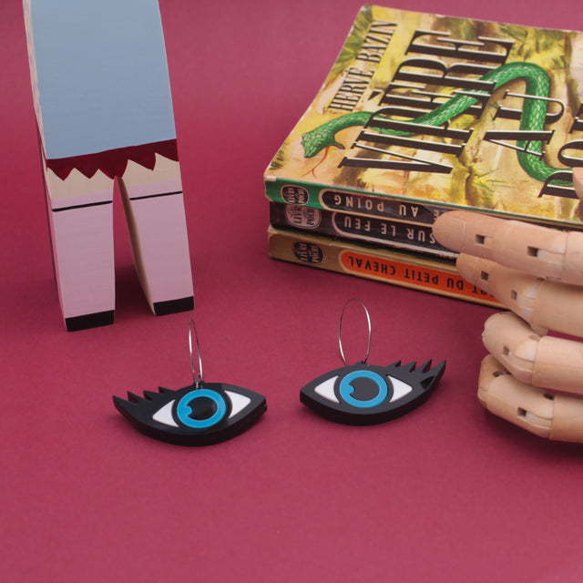 on a red background, at the left a wooden doll, at the right a stack of books and in the center a wooden hand and two evil eye hoops earrings made from black, white and blue laser cut acrylic