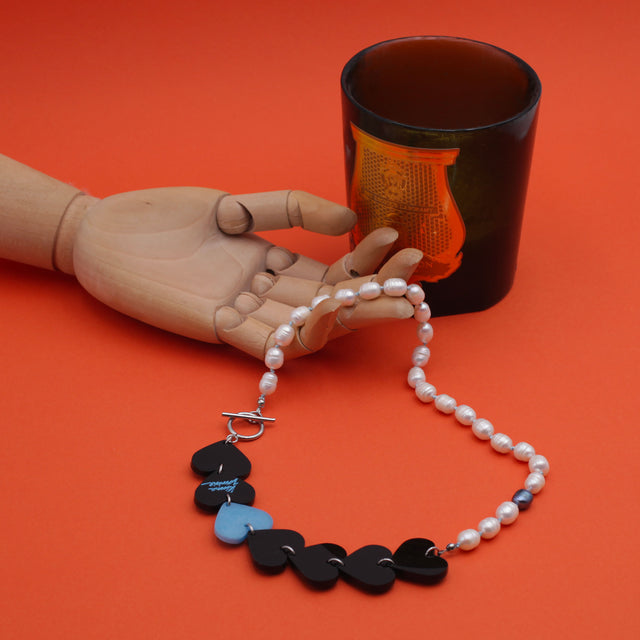 Wooden hand holding a laser-cut and resin black and blue hearts necklace with freshwater pearls and toggle clasp. A dark candle is on the back. On orange background.
