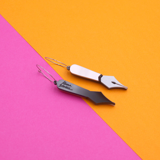 A pair of laser-cut black and pink pearly acrylic pen nib hoops earrings on a pink and orange background. One of the pen nib is on the back and there's a kissmezombie logo engraved. 