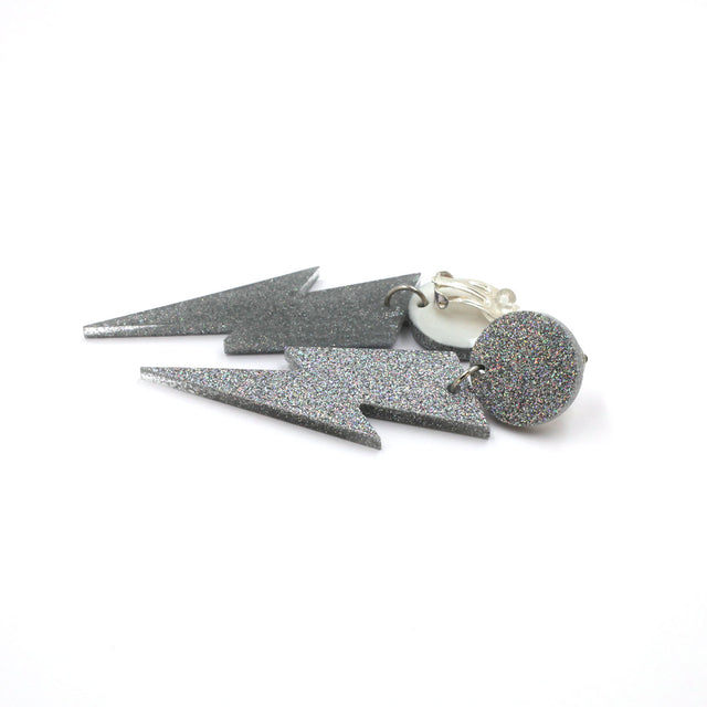 this is a picture of silver holographic glitter bolt and dot dangle earrings on a white background. one is on the back, the clip on are silver on a white dot.