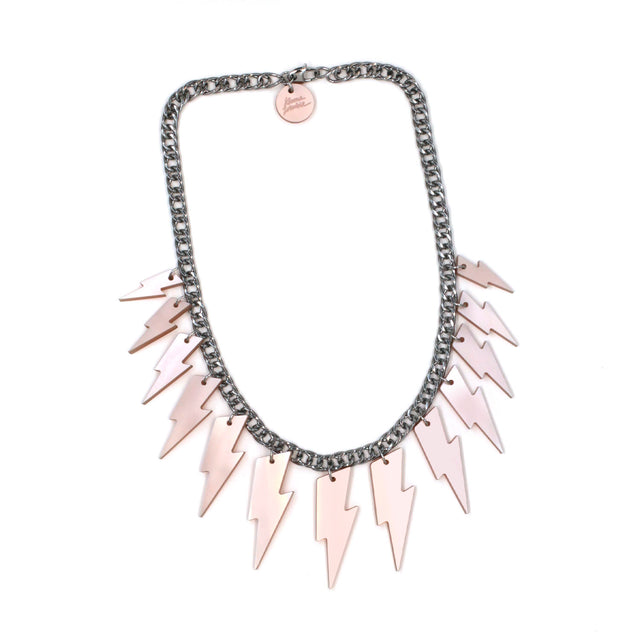 This is a necklace composed of a chunky silver tone curb chain with 13 pieces of pink pearly lightning bolts of graduant size on a white background. on the lobster clasp there is a round charm with the kissmezombie logo engraved on it.