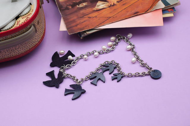 On a purple background, at the center, a laser-cut glittery dark blue acrylic birds and freshwater pearls hanging on a thick curb stainless steel chain with a round charm with the engraved logo "kissmezombie", at the top right corner, there's a tepaz record player and a the left top corner, a stack of vinyls records.