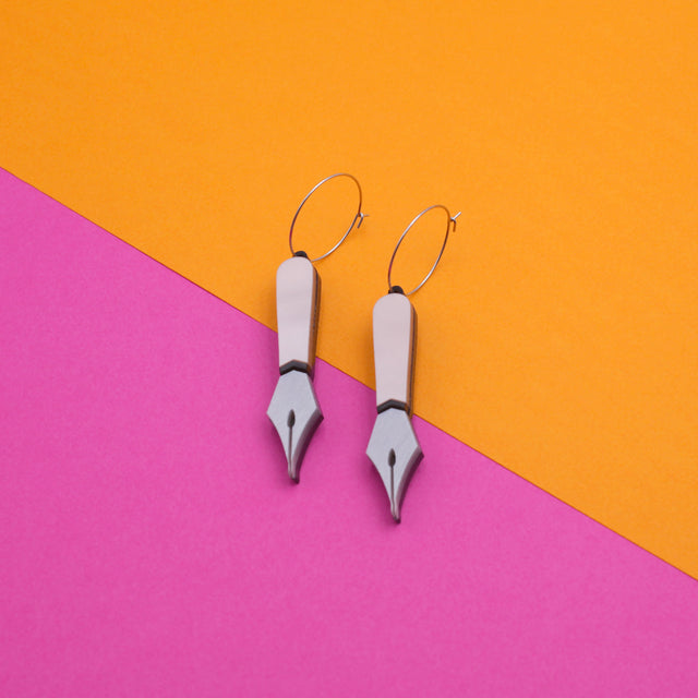 A pair of laser-cut black and pink pearly acrylic pen nib hoops earrings on a pink and orange background.