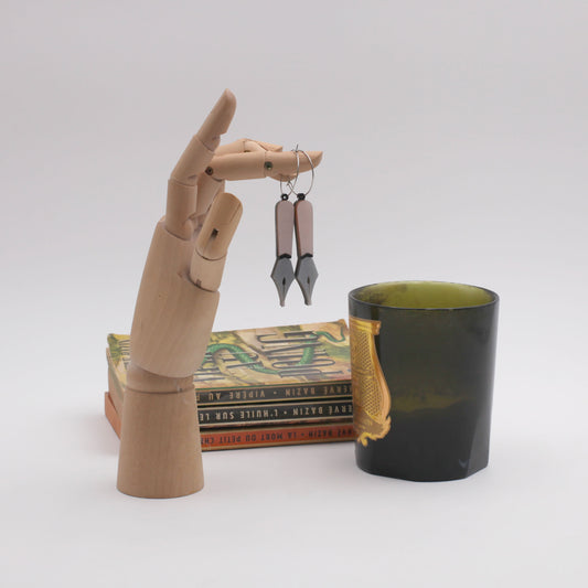 A wooden hand is used to hand a pair of laser-cut black and pink pearly acrylic pen nib hoops earrings. At the left, there's a dark candle with a gold sticker, and in the back a stack of three book on a white background. 