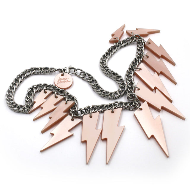 This is a necklace composed of a chunky silver tone curb chain with 13 pieces of pink pearly lightning bolts of graduant size on a white background. on the lobster clasp there is a round charm with the kissmezombie logo engraved on it.