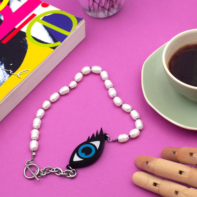 Laser cut evil eye and freshwater pearls necklace on a pink background with a book and a green coffee cup.