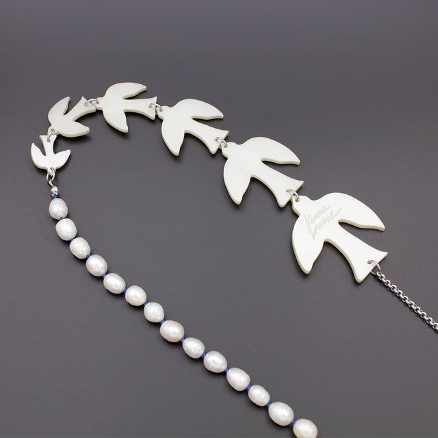 A close up picture of white birds of different sizes and pearls on a sautoir long necklace on a black background. The larger bird is engraved with a kiss me zombie signature logo.