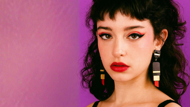 Femme au regard intense portant des boucles d’oreilles statement en forme de tube de rouge à lèvres, en acrylique découpé laser et assemblé à la main. Bijoux faits main à Marseille par Kiss Me Zombie.