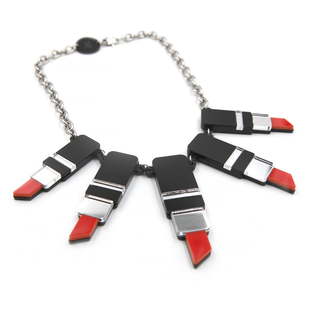 vue rapprochée et de côté un collier plastron tube de rouge à levre en acrylique découpé au laser, noir, rouge et miroir argenté avec une grosse chaine forçat en acier inoxydable sur fond blanc