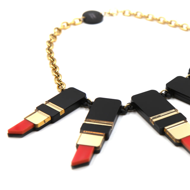 vu de côté du plastron d'un collier composé de tube de rouge à lèvre en acrylique noir, rouge et miroir doré avec une grosse chaîne en acier doré à l'or fin sur fond blanc