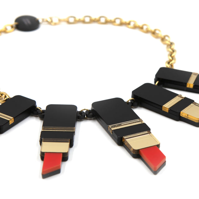 vu de côté du plastron d'un collier composé de tube de rouge à lèvre en acrylique noir, rouge et miroir doré avec une grosse chaîne en acier doré à l'or fin sur fond blanc