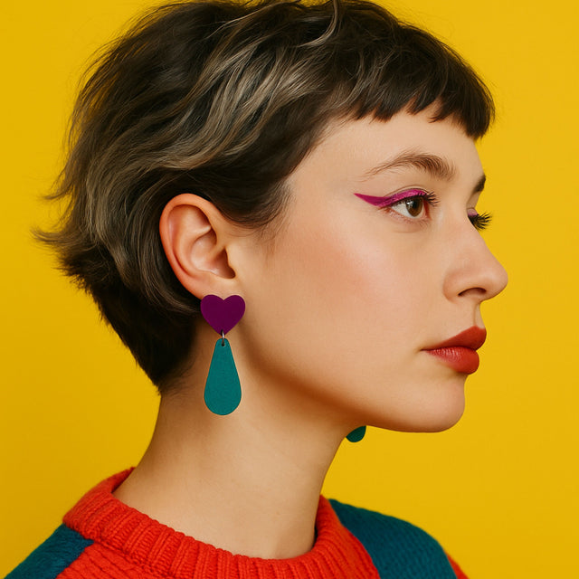 Femme portant des boucles d’oreille statement kiss me zombie composée d’un cœur violet et d’une goutte turquoise, sur fond jaune vibrant. Look coloré, maquillage graphique et coupe courte rebelle.