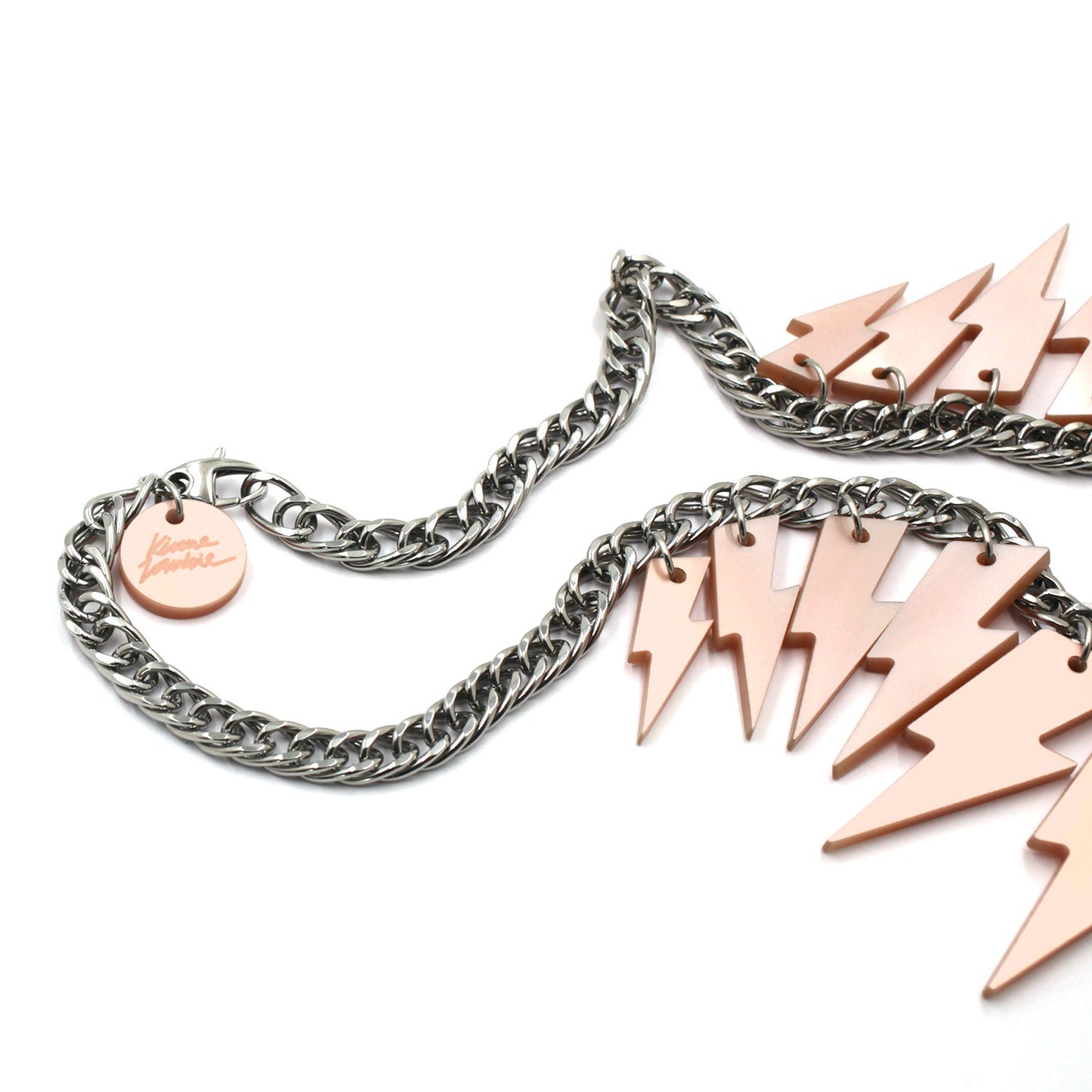 This is a necklace composed of a chunky silver tone curb chain with 13 pieces of pink pearly lightning bolts of graduant size on a white background. on the lobster clasp there is a round charm with the kissmezombie logo engraved on it. 