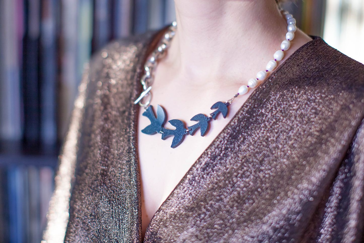 this is a picture of a woman wearing a gold draped top and a statement collar necklace composed by laser cut black birds follow by pearls then a chunky chain. the closure is a T clasp. The background is kind of blurry. it is shelves with vinyl records on them. 