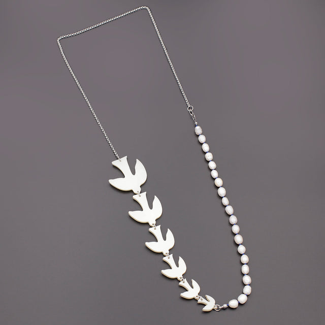 A long necklace is laying on a black background, composed of pearls hand knotted silk thread, 6 birds large to small size and a thin silver tone chain.