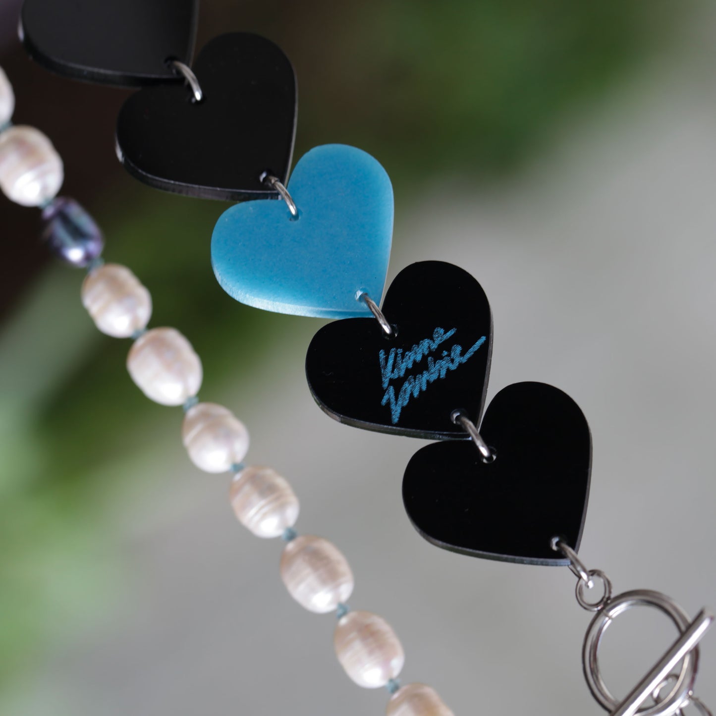 Close-up of a laser-cut and resin black and blue hearts necklace with freshwater pearls and toggle clasp on a blurry background. 