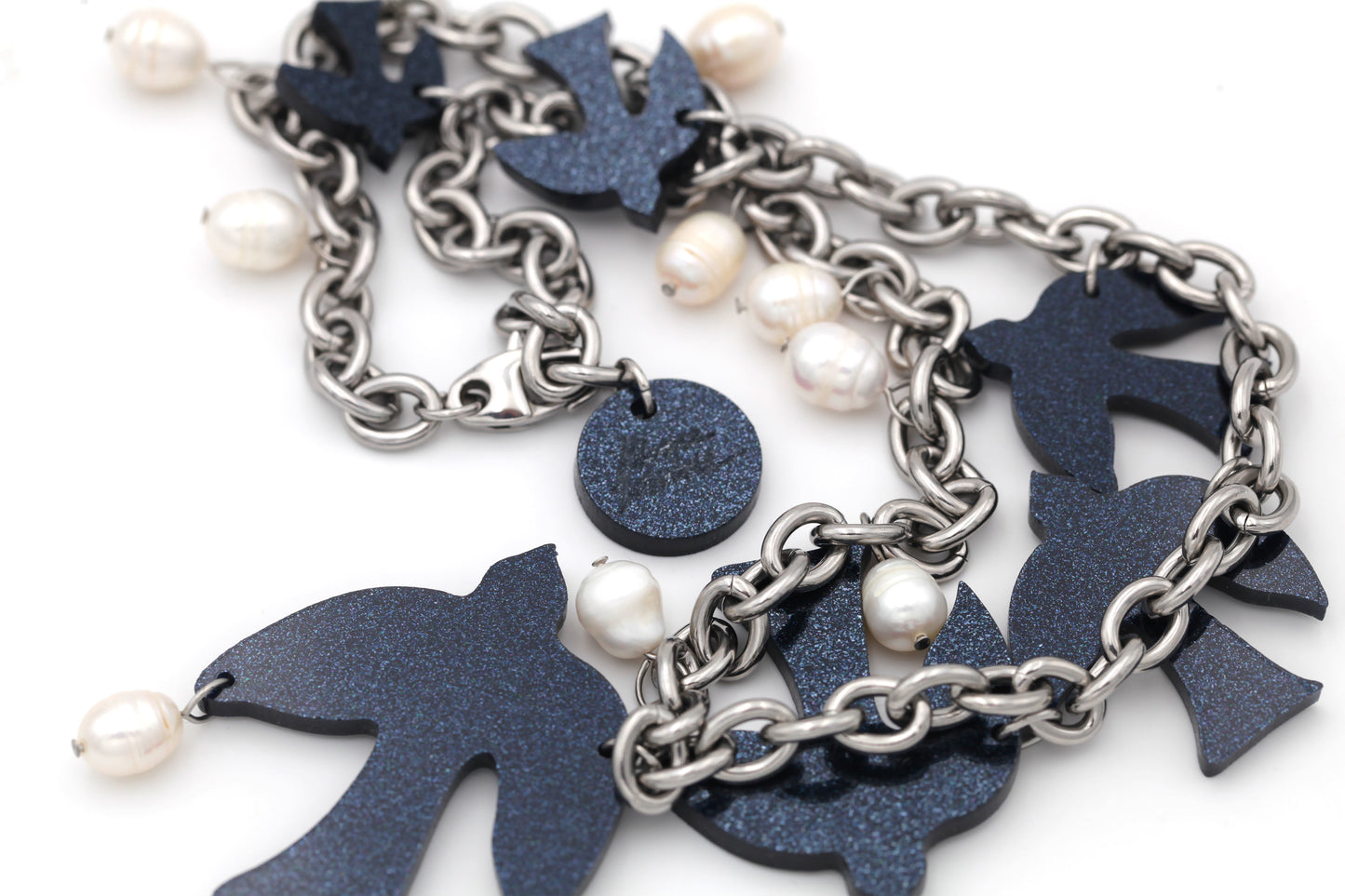 Close-up of dark blue glittery acrylic, laser-cut birds, and white freshwater pearls hanging on a thick steel chain on a white background.