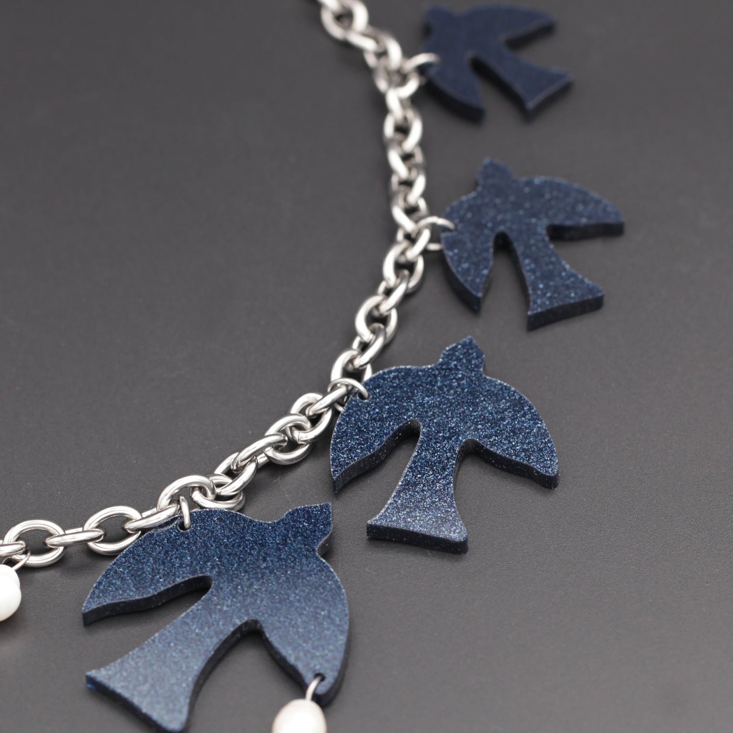 A close-up of a dark blue glittery acrylic, laser-cut birds, and white freshwater pearls hanging on a thick steel chain on a black background.