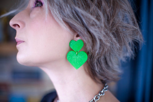 this is the picture of a woman profile. she's dirty blond, her eye are blue and she's wearing bright pink eye shadow. her earrings are bright neon green big heart statement clip on earrings. the background is blurry, dark blue on the right side and a shelves with books on the left side.