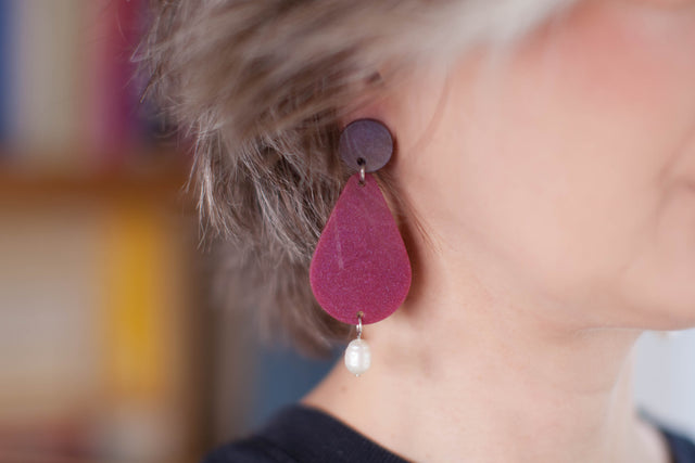 this is a picture of a woman who is wearing statement earrings. we only see half of fer face. the earrings are composed on the top by a purple dot, then a bigger pink shimmering pear and a pearl hanging on the end. the background is blurry and yellowish. 