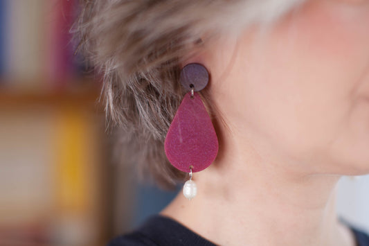 this is a picture of a woman who is wearing statement earrings. we only see half of fer face. the earrings are composed on the top by a purple dot, then a bigger pink shimmering pear and a pearl hanging on the end. the background is blurry and yellowish. 