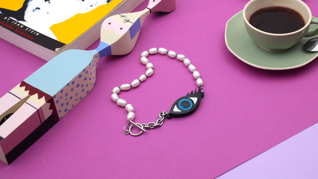 statement, evil eye and pearls necklace, on the right corner, a book, left corner, a green cup of coffee, and a wooden doll on a pink and purple background.  