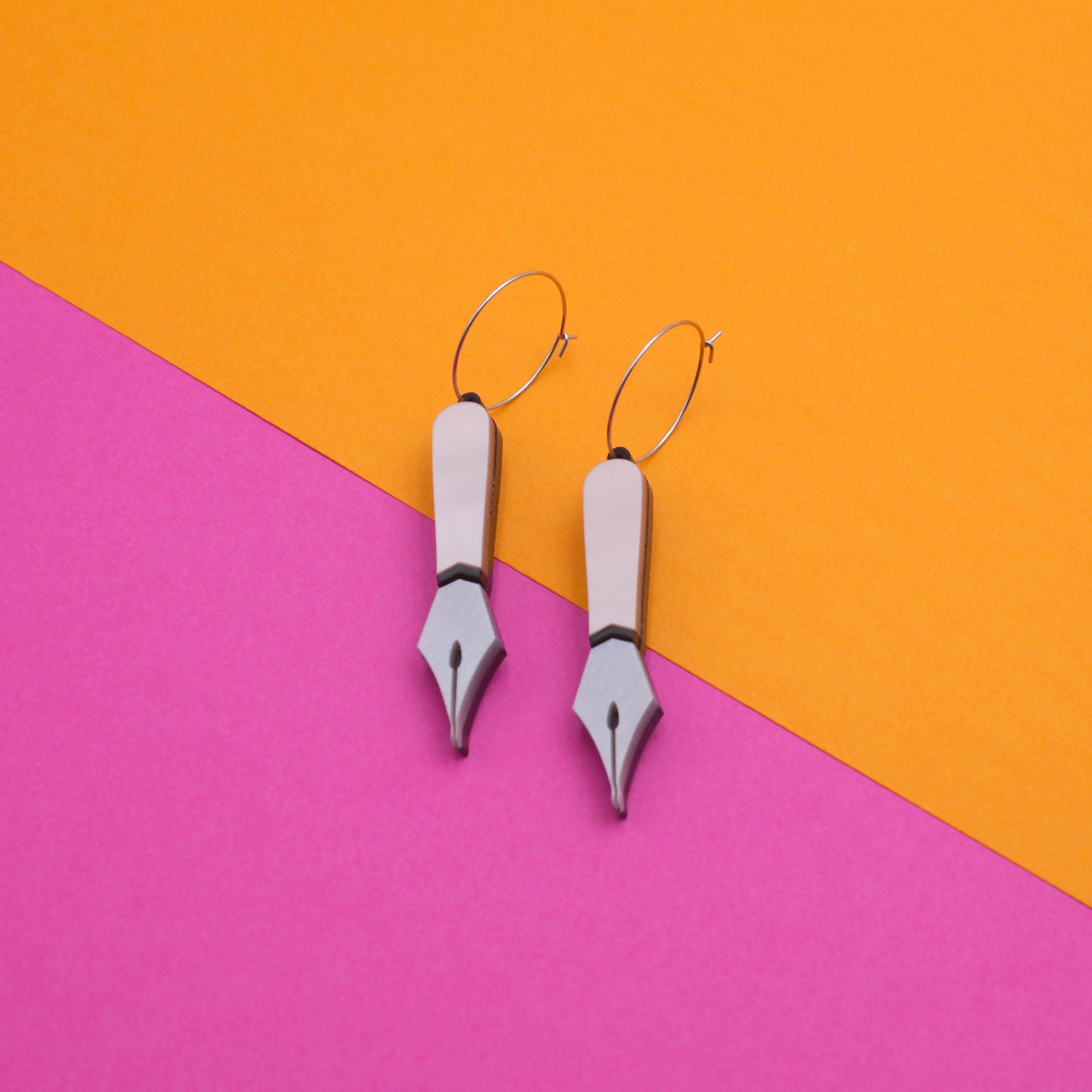 A pair of laser-cut black and pink pearly acrylic pen nib hoops earrings on a pink and orange background. 