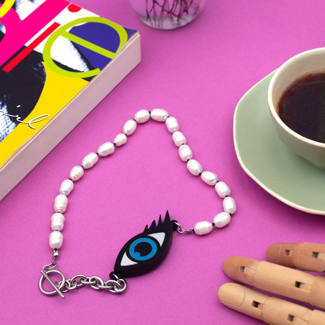 Laser cut evil eye and freshwater pearls necklace on a pink background with a book and a green coffee cup. 