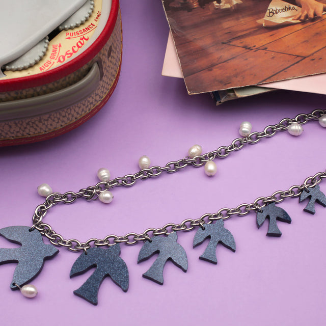 On a purple background, a laser-cut glittery dark blue acrylic birds and freshwater pearls hanging on a thick curb stainless steel chain with a round charm with the engraved logo "kissmezombie", at the top right corner, there's a tepaz record player and a the left top corner, a stack of vinyls records.